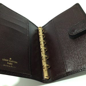 May include: Open brown leather organizer with gold-colored rings and a snap closure. The interior features card slots and the text "LOUIS VUITTON PARIS made in Spain". The organizer is open, displaying its interior.