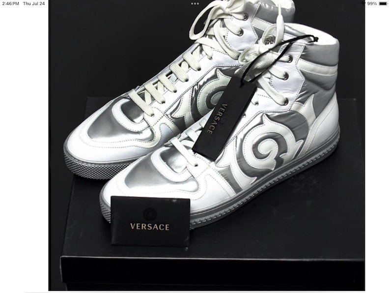 May include: A pair of high-top sneakers in silver and white with decorative swirls. The sneakers have white laces and a silver tongue and heel. A black tag with the brand name "VERSACE" hangs from one shoe. A small black box with the brand name is also present.
