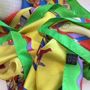 Puede incluir: Un pa&ntilde;uelo de seda con un dise&ntilde;o vibrante. El pa&ntilde;uelo presenta una base amarilla con patrones abstractos coloridos en rojo, azul, morado y verde. Los bordes est&aacute;n ribeteados con un borde verde brillante. El pa&ntilde;uelo es 100% seda.