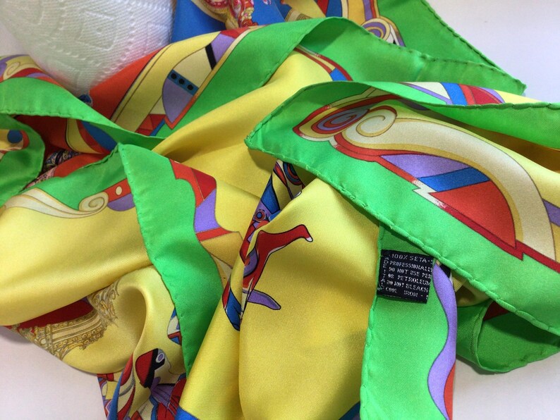 Puede incluir: Un pa&ntilde;uelo de seda con un dise&ntilde;o vibrante. El pa&ntilde;uelo presenta una base amarilla con patrones abstractos coloridos en rojo, azul, morado y verde. Los bordes est&aacute;n ribeteados con un borde verde brillante. El pa&ntilde;uelo es 100% seda.