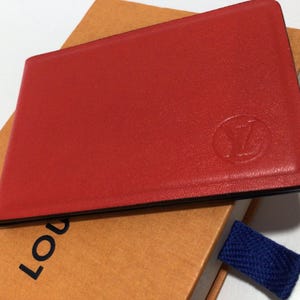 May include: A rectangular red leather card holder featuring a circular embossed logo on the front surface. The card holder is positioned on top of a tan box with the partial black text "LOU" visible and a small blue fabric pull tab attached.