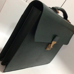 May include: A dark forest green briefcase with a textured surface, a top handle, and a gold-toned metal clasp closure. The bag features a structured rectangular shape with a flap cover that secures over the front compartment with a metallic latch.