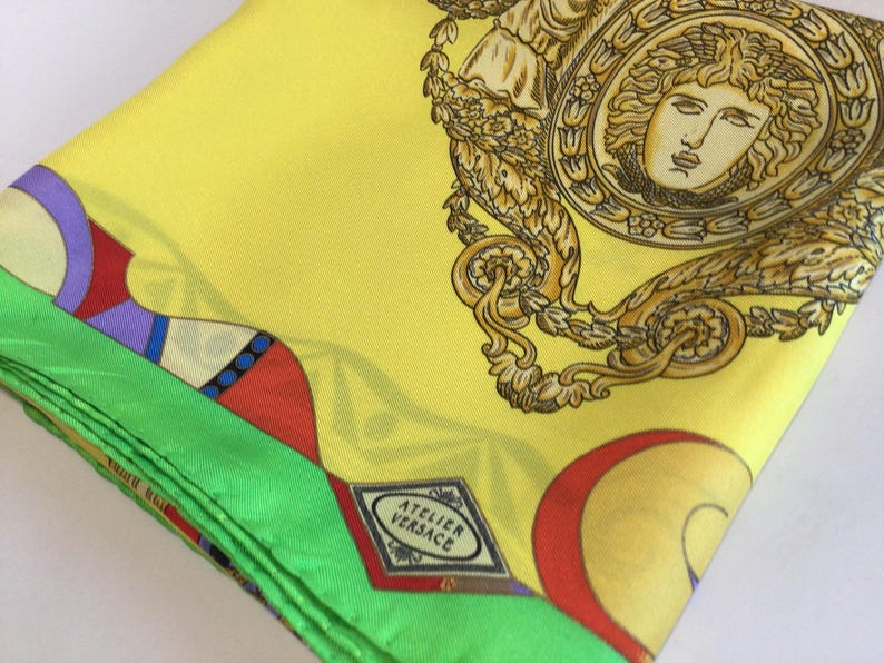 Puede incluir: Un pa&ntilde;uelo de seda amarillo doblado con un borde verde y patrones geom&eacute;tricos coloridos. El pa&ntilde;uelo presenta un dise&ntilde;o de cabeza de Medusa dorada y el logotipo "ATELIER VERSACE".