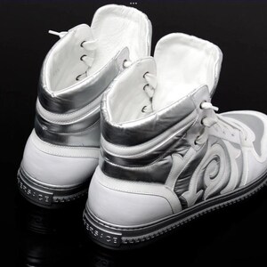 May include: White high-top sneakers with silver accents. The sneakers feature a unique design on the side and silver trim. The brand name is visible on the sole. The shoes are displayed on a black surface.