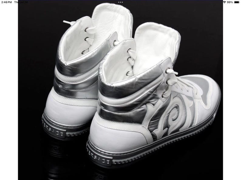 May include: White high-top sneakers with silver accents. The sneakers feature a unique design on the side and silver trim. The brand name is visible on the sole. The shoes are displayed on a black surface.