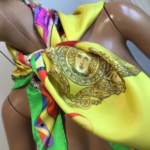 Puede incluir: Un pa&ntilde;uelo de seda amarillo brillante con un dise&ntilde;o de cabeza de Medusa dorada y un patr&oacute;n geom&eacute;trico colorido. El pa&ntilde;uelo est&aacute; drapeado alrededor del cuello de un maniqu&iacute;, mostrando sus colores vibrantes y detalles intrincados.