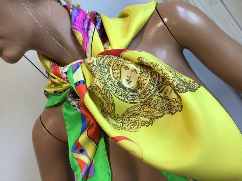 Puede incluir: Un pa&ntilde;uelo de seda amarillo brillante con un dise&ntilde;o de cabeza de Medusa dorada y un patr&oacute;n geom&eacute;trico colorido. El pa&ntilde;uelo est&aacute; drapeado alrededor del cuello de un maniqu&iacute;, mostrando sus colores vibrantes y detalles intrincados.