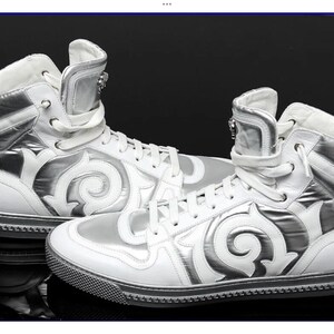 May include: Pair of white high-top sneakers with silver accents. The sneakers feature a unique design with a swirling pattern on the sides and a zipper detail on the tongue. The soles are black with a textured pattern.