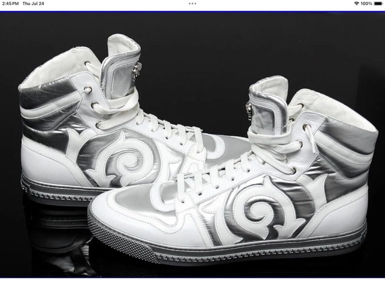 May include: Pair of white high-top sneakers with silver accents. The sneakers feature a unique design with a swirling pattern on the sides and a zipper detail on the tongue. The soles are black with a textured pattern.