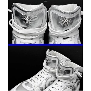 May include: High-top white sneakers with silver accents. The sneakers feature a silver panel on the upper and a Medusa head emblem. White laces and a padded collar complete the design. The shoes are set against a black background.