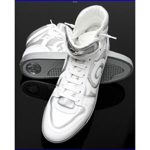 May include: White high-top sneakers with silver accents. The sneakers feature a lace-up design, a padded collar, and a silver decorative pattern on the sides. The soles are gray and textured. The shoes are displayed on a black surface.