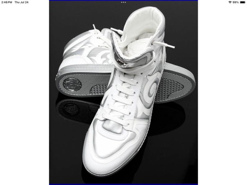 May include: White high-top sneakers with silver accents. The sneakers feature a lace-up design, a padded collar, and a silver decorative pattern on the sides. The soles are gray and textured. The shoes are displayed on a black surface.
