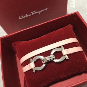 May include: A light pink leather bracelet with a silver-tone clasp, presented on a red velvet cushion inside a red box. The box lid is open, revealing the bracelet and the brand name, Salvatore Ferragamo.