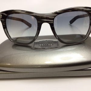 May include: Sunglasses with a dark gray, patterned frame and gradient lenses. The sunglasses rest on a matching gray case with the brand name "Oliver Peoples" embossed on it. The overall design is sleek and modern.