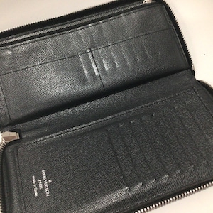May include: Open black leather wallet with multiple card slots and compartments. The wallet has a full zipper closure and a silver zipper pull. The interior is black with a textured finish. The brand name is visible.