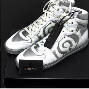 May include: A pair of high-top sneakers in silver and white with decorative swirls. The sneakers have white laces and a silver tongue and heel. A black tag with the brand name "VERSACE" hangs from one shoe.
