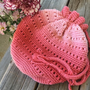 Könnte beinhalten: Eine rosa gehäkelte Kordeltasche mit einem Farbverlauf von Dunkelrosa zu Hellrosa. Die Tasche hat an jeder Seite einen Quasten und ist aus einem dicken Garn gefertigt.