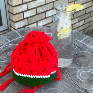 Könnte beinhalten: Eine gehäkelte Wassermelonen-Tasche mit grünen, weißen und roten Streifen. Die Tasche hat einen Kordelverschluss und liegt auf einer grauen Steinoberfläche.