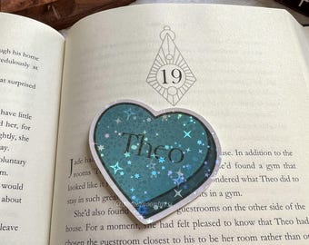 Puede incluir: Una pegatina en forma de corazón azul verdoso con el nombre "Theo" en letras blancas y estrellas plateadas brillantes. La pegatina está colocada en una página de un libro con el número "19" impreso en ella.
