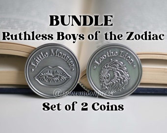 Puede incluir: Un conjunto de dos monedas de metal plateadas con diseños grabados. La primera moneda presenta un labio rojo con colmillos y el texto "Little Monster". La segunda moneda presenta una cabeza de león y el texto "Leo the Lion". El texto "Bundle Ruthless Boys of the Zodiac Set of 2 Coins" también es visible.