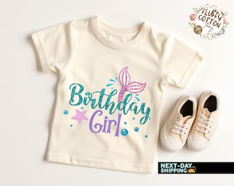 May include: A cream-colored t-shirt with the words "Birthday Girl" in teal glitter script, a purple mermaid tail, and a starfish. The shirt is paired with beige sneakers. The shirt is likely for a child's birthday.