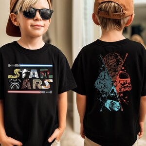 May include: Black t-shirt featuring the text "STAR WARS" in a colorful, stylized font with lightsabers. The back of the shirt displays a graphic with Star Wars characters in red, blue, and brown.