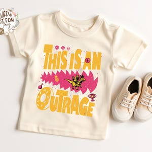 May include: Cream-colored toddler t-shirt with the words "THIS IS AN OUTRAGE" in yellow, with a pink and yellow graphic of a dinosaur and other elements. A pair of beige canvas sneakers are next to the shirt.