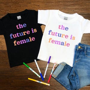 May include: Two t-shirts with the text "the future is female" printed on them in rainbow colors. The shirts are on a wooden surface with a pair of blue jeans and colorful markers.