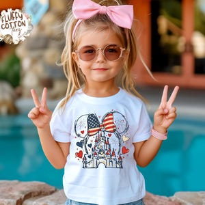 May include: A young child wearing a white t-shirt with a colorful graphic of a castle, hearts, and American flag-themed mouse ears. The child is wearing sunglasses and a pink bow. The child is giving the peace sign.