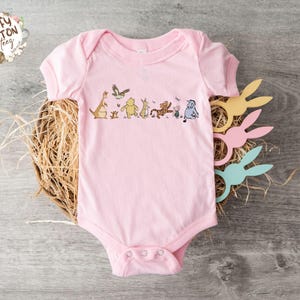 May include: A pink baby onesie with a graphic of Winnie the Pooh and his friends, including Tigger, Piglet, Eeyore, and Kanga, walking in a line. The onesie has a snap closure at the crotch.