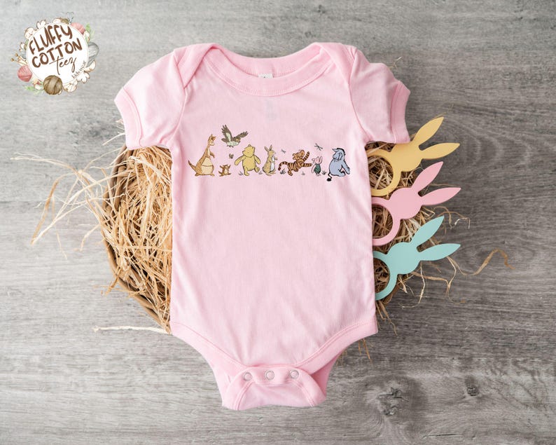 May include: A pink baby onesie with a graphic of Winnie the Pooh and his friends, including Tigger, Piglet, Eeyore, and Kanga, walking in a line. The onesie has a snap closure at the crotch.