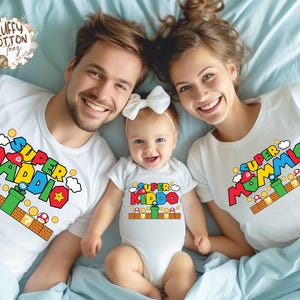 May include: A family wearing matching white t-shirts and a onesie with colorful "Super Mario" inspired graphics. The shirts say "Super Daddio" and "Super Mommio", while the onesie says "Super Kiddo". The family is lying on a light blue bedspread.
