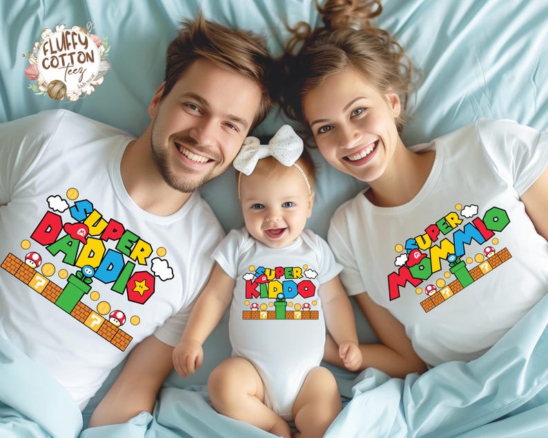 May include: A family wearing matching white t-shirts and a onesie with colorful "Super Mario" inspired graphics. The shirts say "Super Daddio" and "Super Mommio", while the onesie says "Super Kiddo". The family is lying on a light blue bedspread.