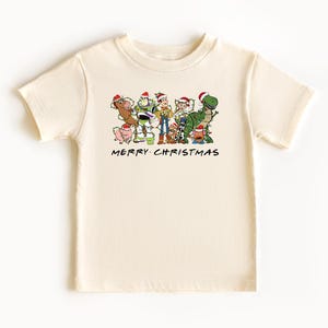 May include: A cream colored t-shirt with a Christmas themed graphic of the Toy Story characters wearing Santa hats. The text "Merry Christmas" is printed below the graphic.