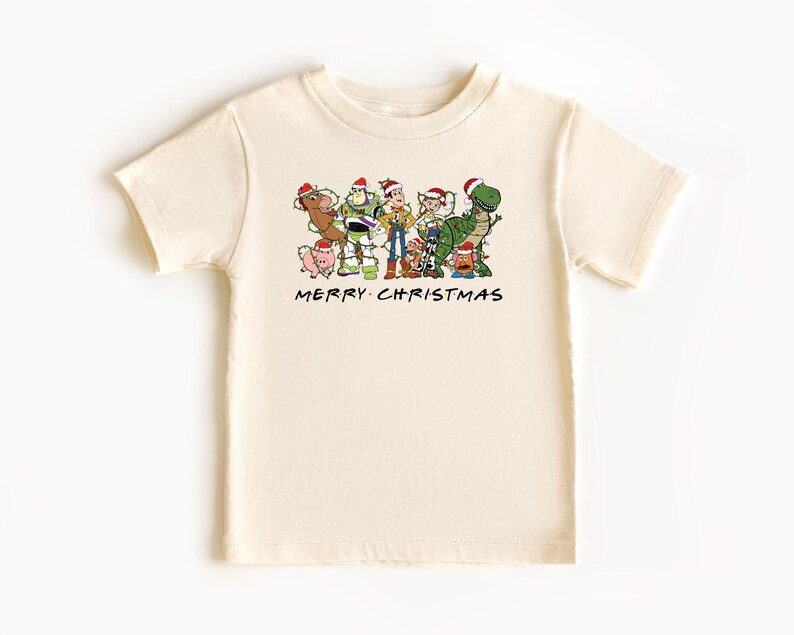 May include: A cream colored t-shirt with a Christmas themed graphic of the Toy Story characters wearing Santa hats. The text "Merry Christmas" is printed below the graphic.