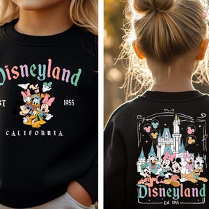 May include: Black sweatshirt with "Disneyland" in a colorful script, featuring Mickey Mouse, Donald Duck, and other Disney characters. The sweatshirt also has "EST 1955" and "CALIFORNIA" printed on it.