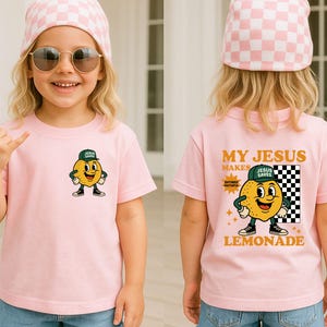 K&ouml;nnte beinhalten: Rosa kurz&auml;rmeliges T-Shirt mit einem Cartoon-Zitronen-Charakter, der eine gr&uuml;ne Kappe mit der Aufschrift "Jesus Saves" tr&auml;gt. Auf der R&uuml;ckseite des Shirts steht "My Jesus Makes Lemonade". Das Kind tr&auml;gt eine rosa-wei&szlig; karierte M&uuml;tze und eine Sonnenbrille.