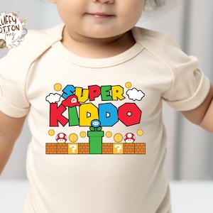 May include: A cream-colored baby onesie featuring the text "SUPER KIDDO" in colorful, video game-inspired lettering. The design includes a red hat, clouds, coins, and a green pipe with a flower. The onesie has short sleeves.