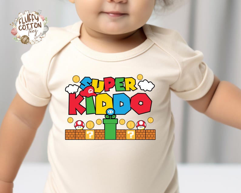May include: A cream-colored baby onesie featuring the text "SUPER KIDDO" in colorful, video game-inspired lettering. The design includes a red hat, clouds, coins, and a green pipe with a flower. The onesie has short sleeves.