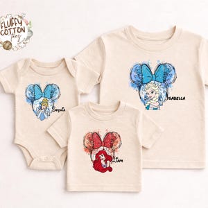 以下が含まれることがあります： 3枚のベージュ色のTシャツと、ディズニーのプリンセスデザインのボディスーツ。各衣類には、プリンセスの画像と名前が付いたミッキーマウスの耳のシルエットがあります。デザインには、シンデレラ、エルサ、アリエルが含まれています。