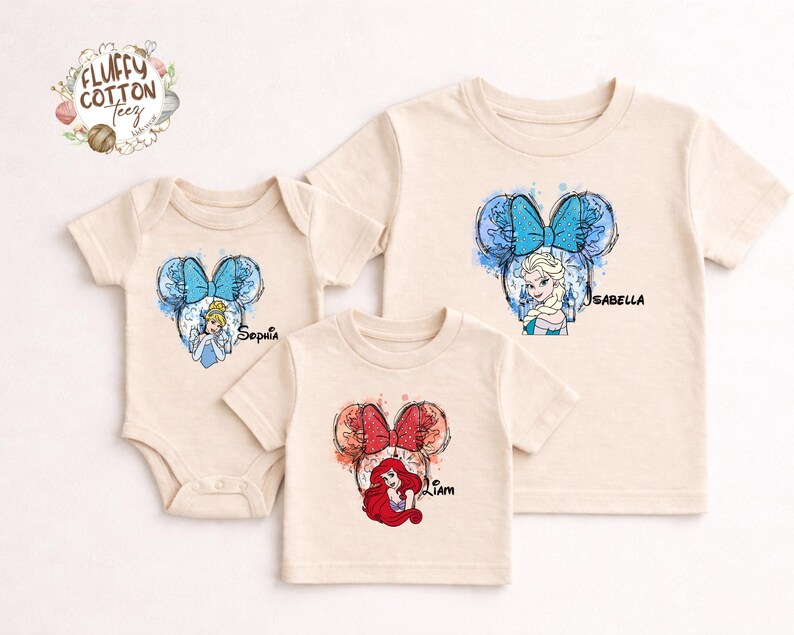 以下が含まれることがあります： 3枚のベージュ色のTシャツと、ディズニーのプリンセスデザインのボディスーツ。各衣類には、プリンセスの画像と名前が付いたミッキーマウスの耳のシルエットがあります。デザインには、シンデレラ、エルサ、アリエルが含まれています。