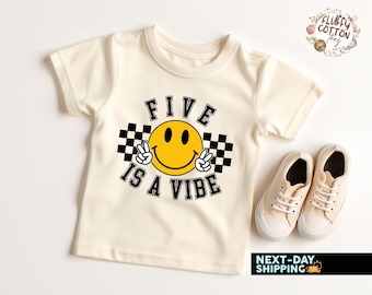 May include: A cream colored t-shirt with a black and white checkered design and a yellow smiley face with peace signs. The text "FIVE IS A VIBE" is printed in black.