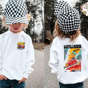 May include: A white sweatshirt with a Lightning McQueen design. The front has the number "95" and a race car graphic, and the back features a colourful "Lightning McQueen" poster from the "Cars" film. The child is wearing a black and white chequered cap.