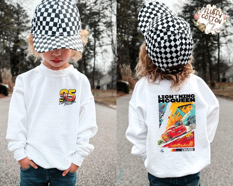 May include: A white sweatshirt with a Lightning McQueen design. The front has the number "95" and a race car graphic, and the back features a colourful "Lightning McQueen" poster from the "Cars" film. The child is wearing a black and white chequered cap.