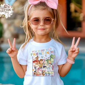 Op de afbeelding: Een wit T-shirt met de tekst "YOU'VE GOT A FRIEND IN ME" en een kleurrijke afbeelding van Toy Story-personages. Het meisje draagt een roze strik, een zonnebril en een roze armband.