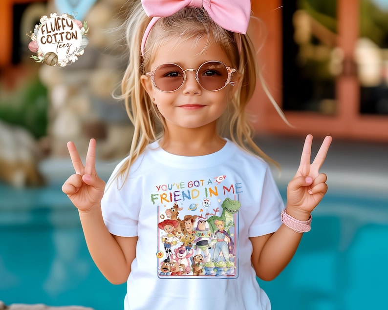Op de afbeelding: Een wit T-shirt met de tekst "YOU'VE GOT A FRIEND IN ME" en een kleurrijke afbeelding van Toy Story-personages. Het meisje draagt een roze strik, een zonnebril en een roze armband.