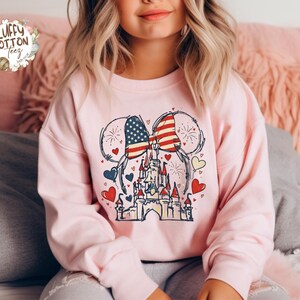 May include: A light pink sweatshirt featuring a graphic of a castle with an American flag bow and mouse ears. The design includes red, white, and blue hearts and fireworks. The sweatshirt has long sleeves.