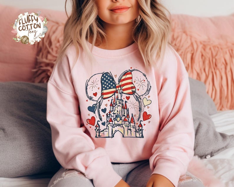 May include: A light pink sweatshirt featuring a graphic of a castle with an American flag bow and mouse ears. The design includes red, white, and blue hearts and fireworks. The sweatshirt has long sleeves.