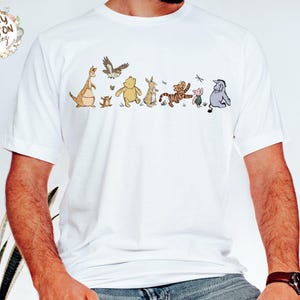 May include: White t-shirt with a line of Winnie the Pooh characters, including Tigger, Piglet, Eeyore, and Roo. The characters are printed in brown, yellow, and pink.