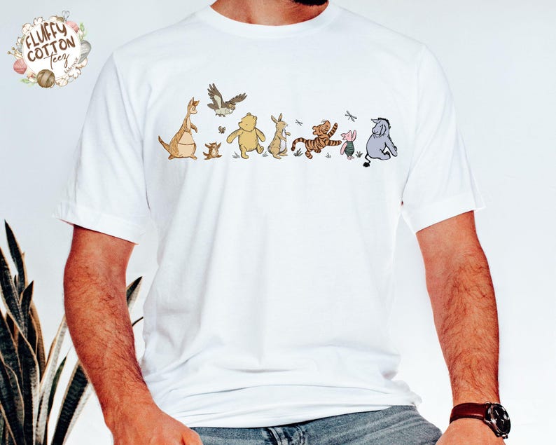 May include: White t-shirt with a line of Winnie the Pooh characters, including Tigger, Piglet, Eeyore, and Roo. The characters are printed in brown, yellow, and pink.